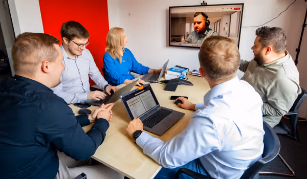 Pięć osób siedzi przy stole konferencyjnym, korzystając z laptopów, a na ekranie telewizora prowadzą wideokonferencję z mężczyzną.