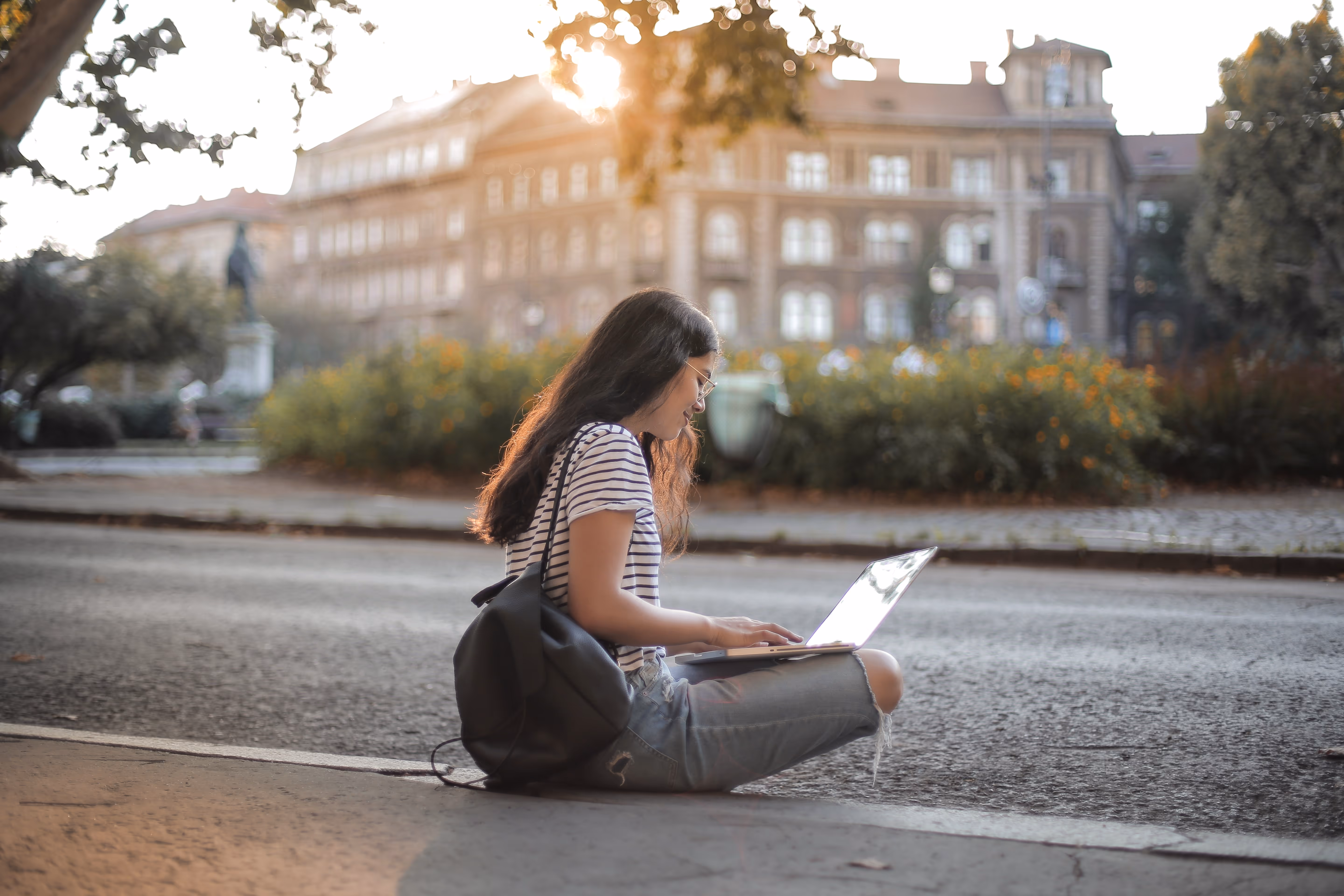 Młoda kobieta siedzi na chodniku na zewnątrz, pracując na laptopie, z plecakiem i budynkami w tle oświetlonymi światłem słonecznym.