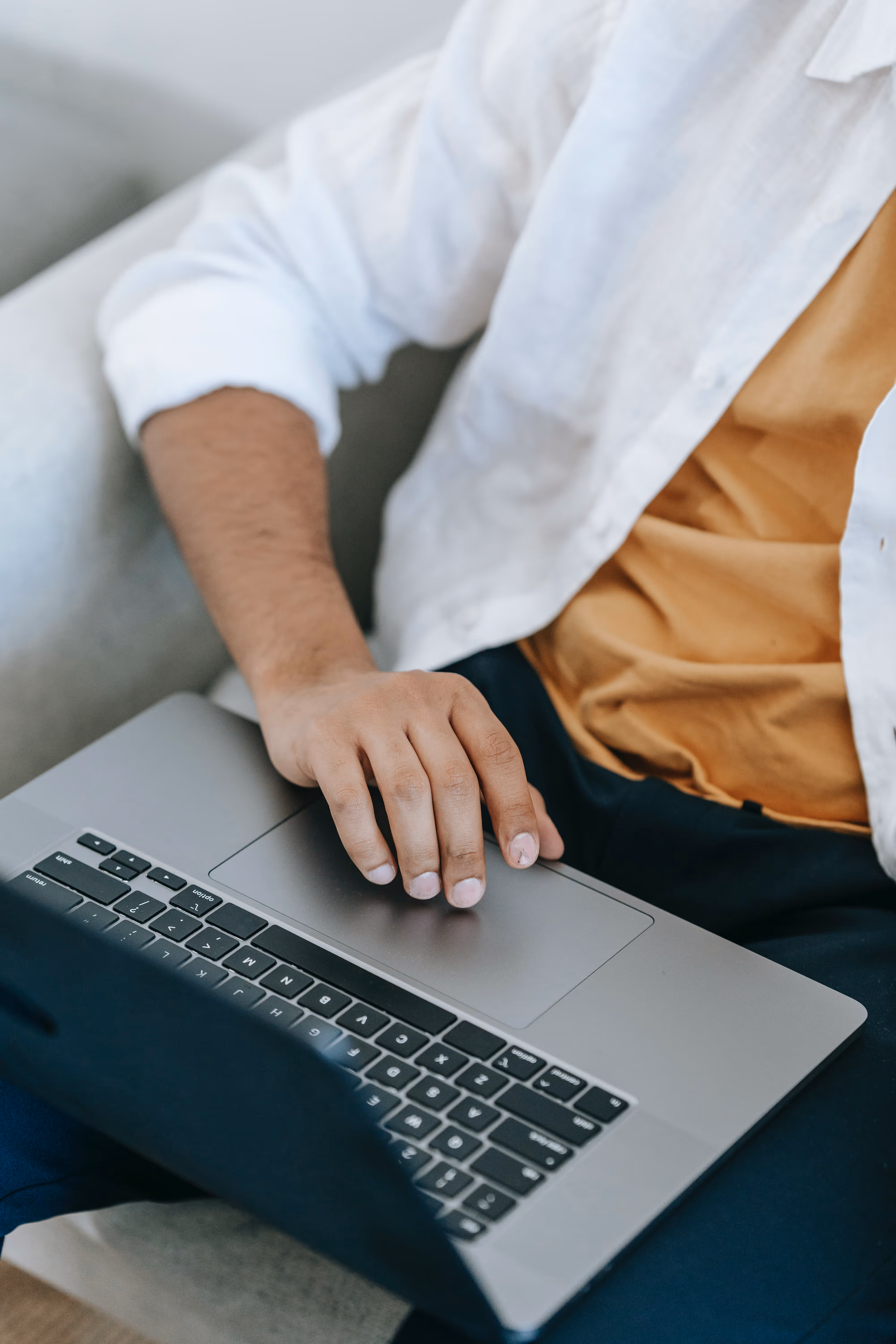 Ręka osoby korzystającej z laptopa, leżącej na kolanach, ubranej w białą koszulę i pomarańczową koszulkę.