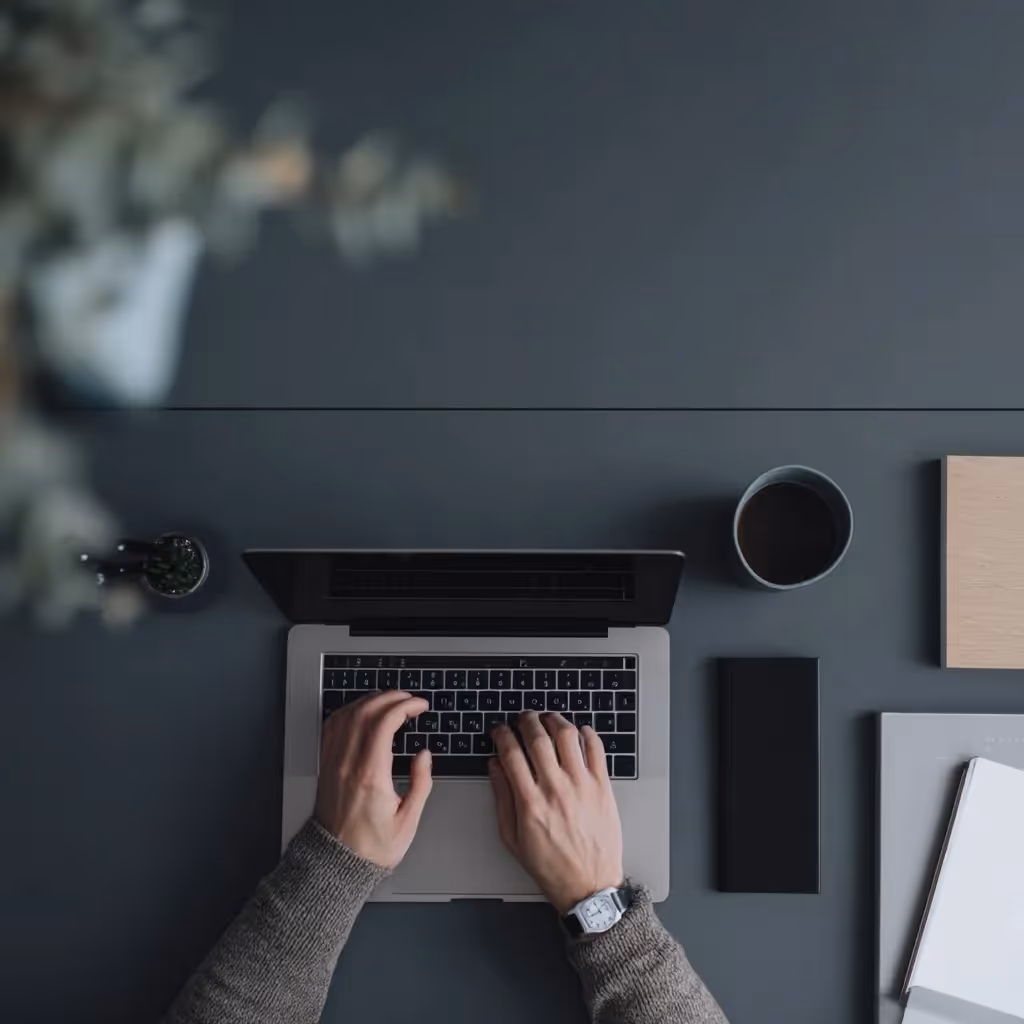 Dłoń osoby piszącej na laptopie umieszczonym na ciemnym biurku obok małej rośliny doniczkowej.