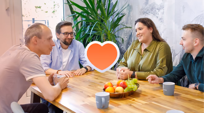 Cztery osoby rozmawiają przy stole z owocami i kubkami, na środku ikona serca.