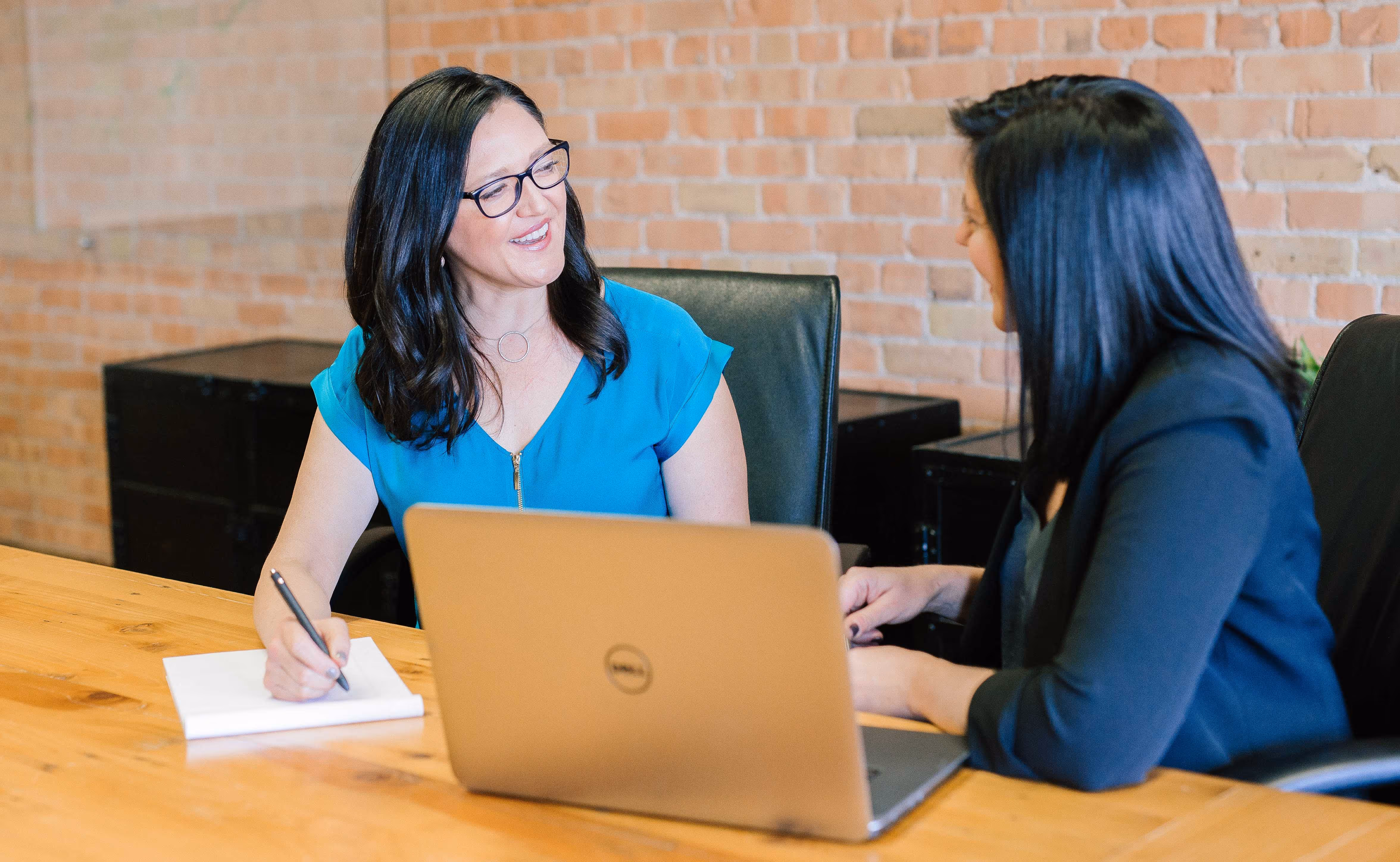 Two people sitting next to each other talking. One is on a laptop and the other is writing notes.