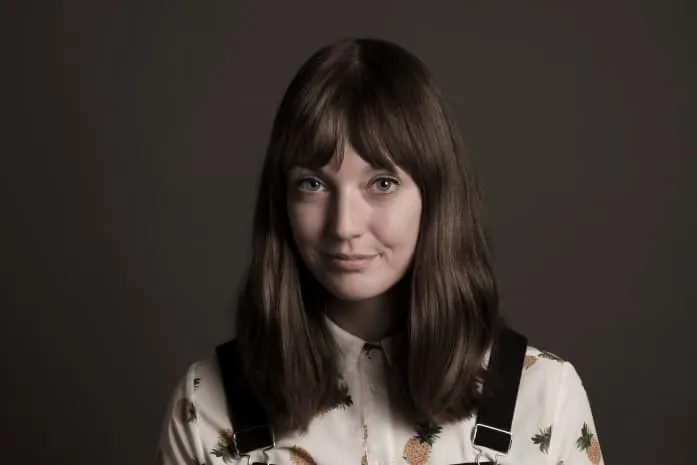 Professionelt virksomhedsportræt af ung kvinde med glat brunt hår og pandehår, iført hvid skjorte med ananasprint og sorte seler mod mørk baggrund – ideelt til medarbejderprofil, LinkedIn-portræt og corporate branding.