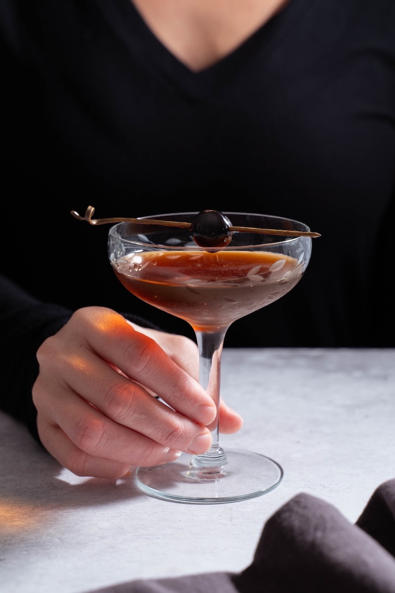 Person holding the stem of a cocktail glass filled with a Black Manhattan drink