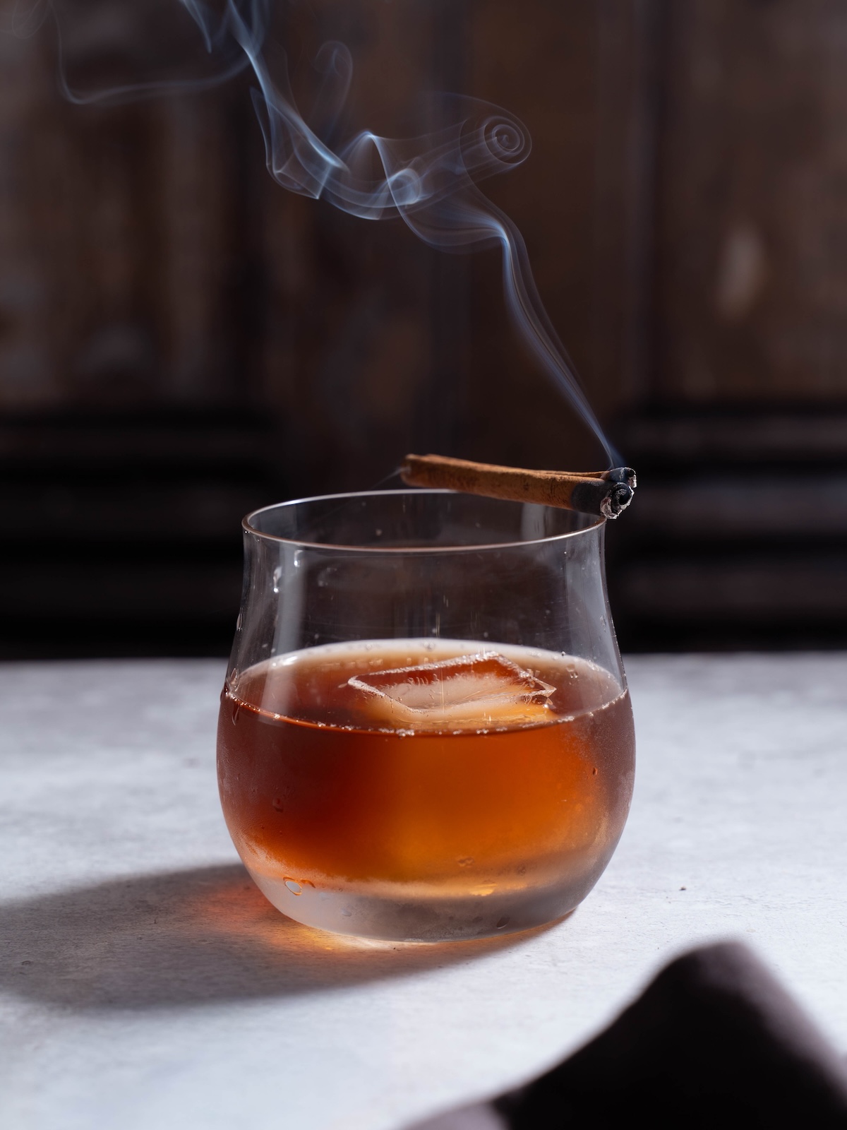 A cinnamon stick smoking on the side of a glass of cold whiskey