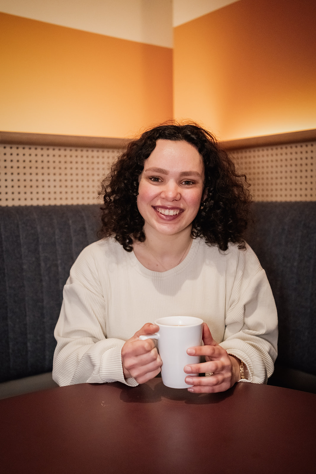 Tara Pigott, Graphic & Brand Designer, smiling and holding coffee mug sat at a table