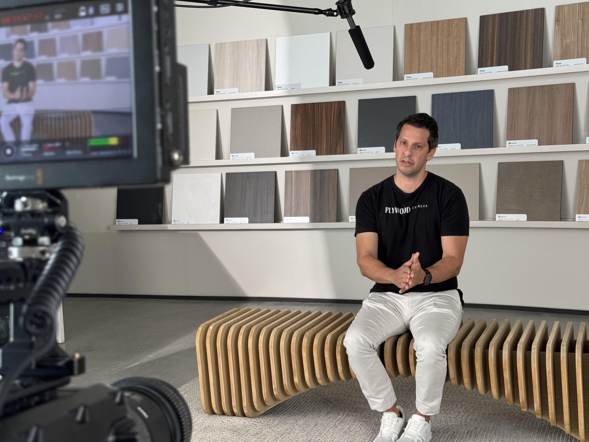 Plywood Express BTS — subject at interview facing camera, Plywood Express shirt, full wood sample wall