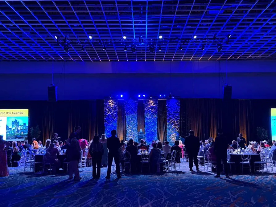 Guests mingling at The Faine House Gala under blue light with a stage screen