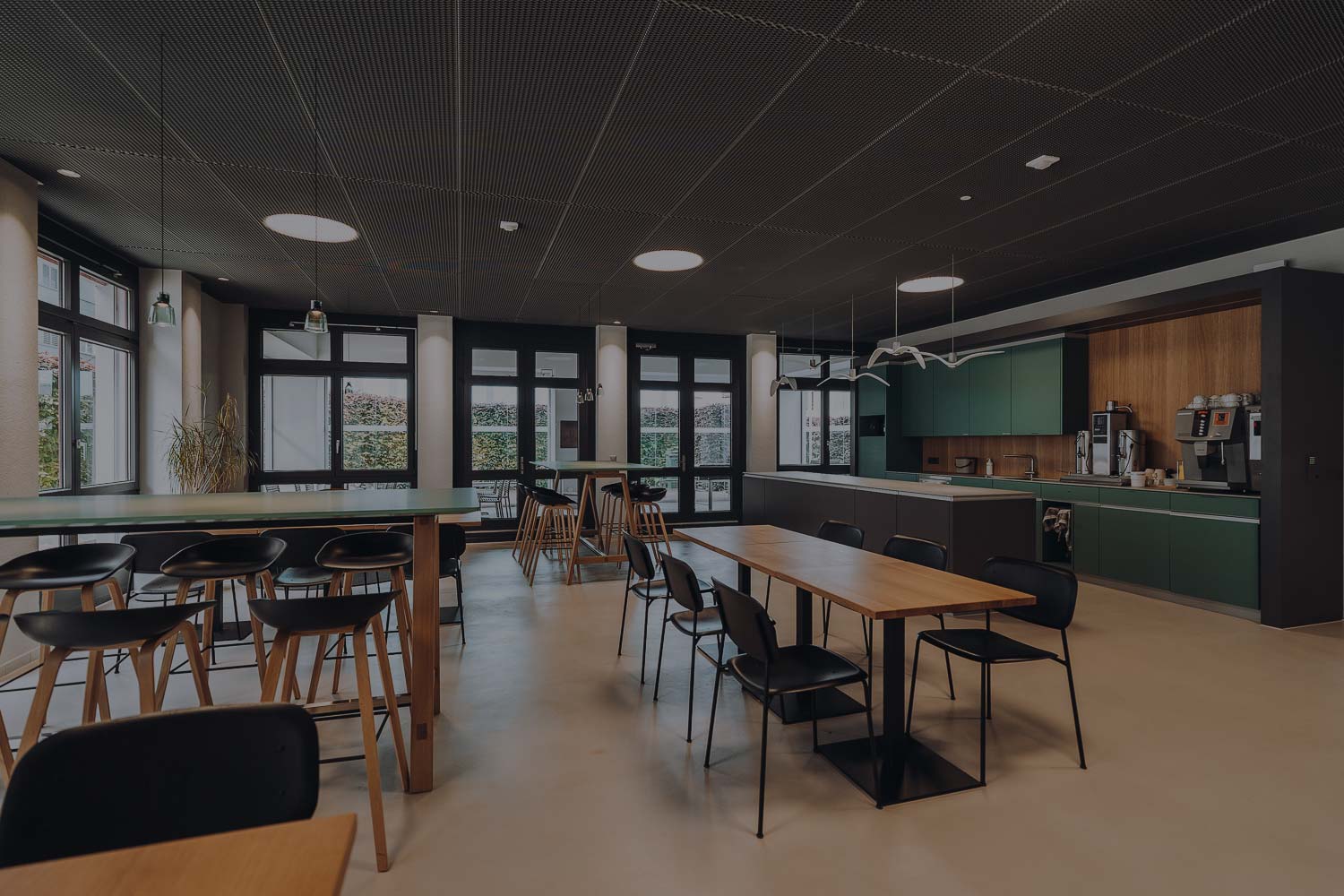 Modern kitchen and dining area with wooden tables, black chairs, green cabinets, and coffee machines along the back wall.