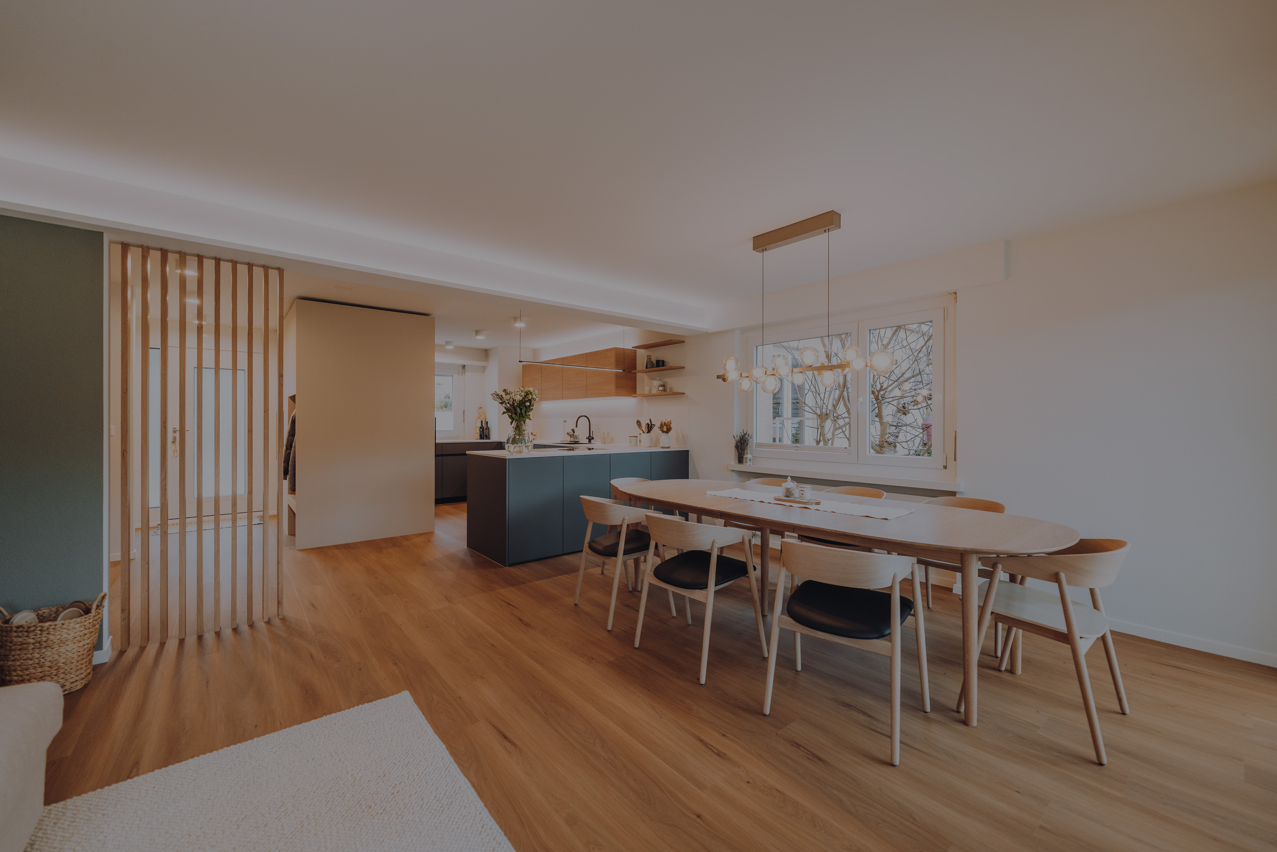 Modern dining room with wooden table, six chairs, hanging light fixture, and kitchen area in the background.