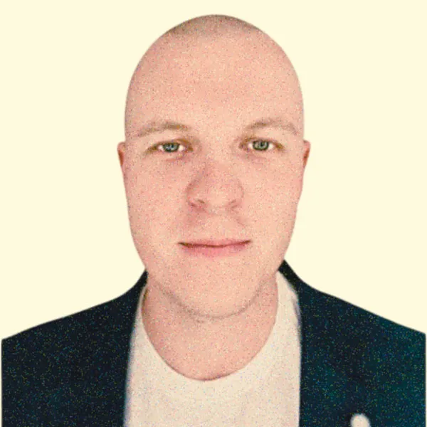 Portrait of a bald man with light skin wearing a dark jacket and white shirt against a pale background.