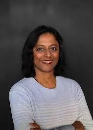 Smiling woman with medium-dark skin and shoulder-length black hair wearing a light gray sweater against a dark background.