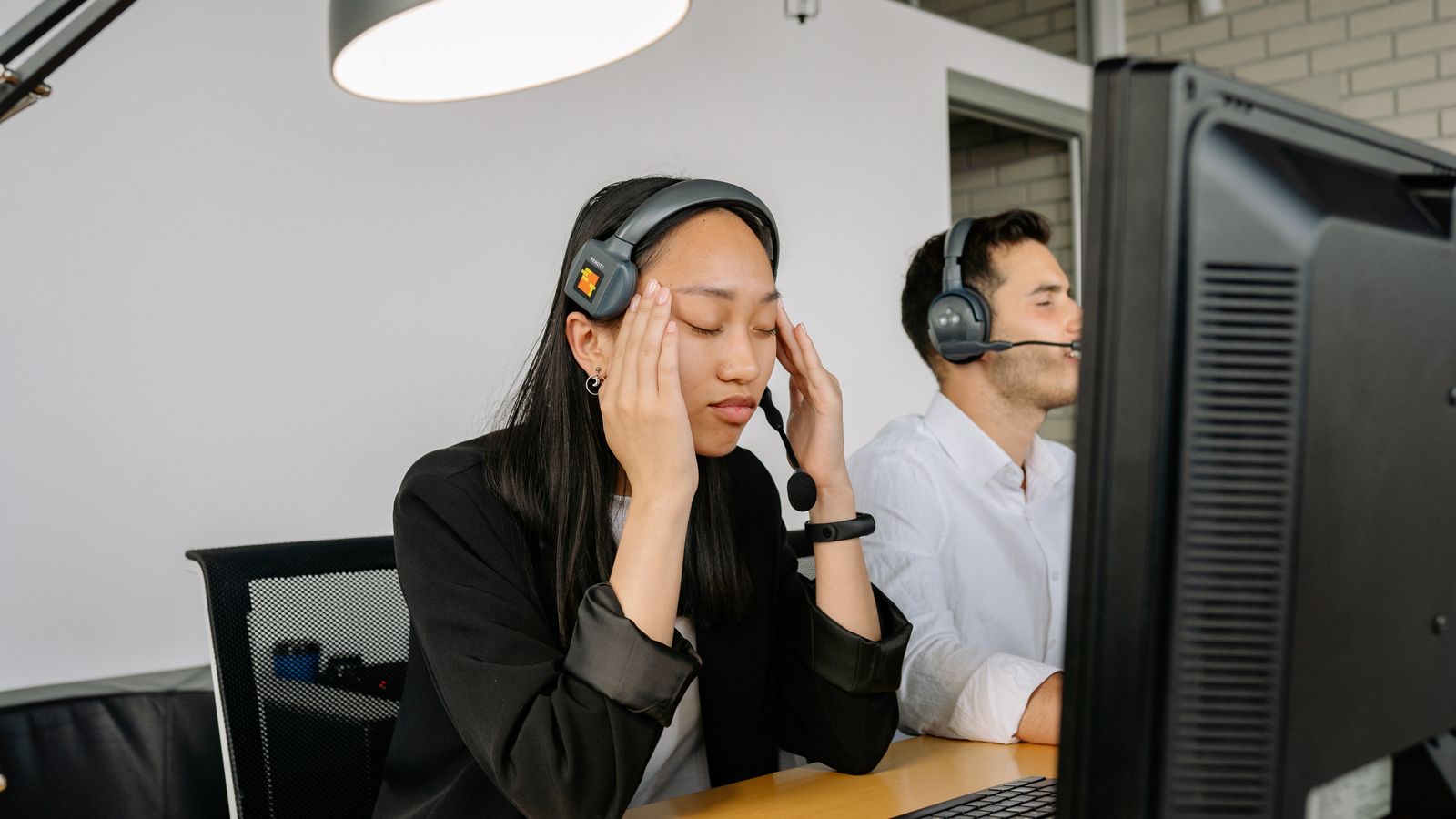 Asian woman at call center desk looking stressed, eyes closed