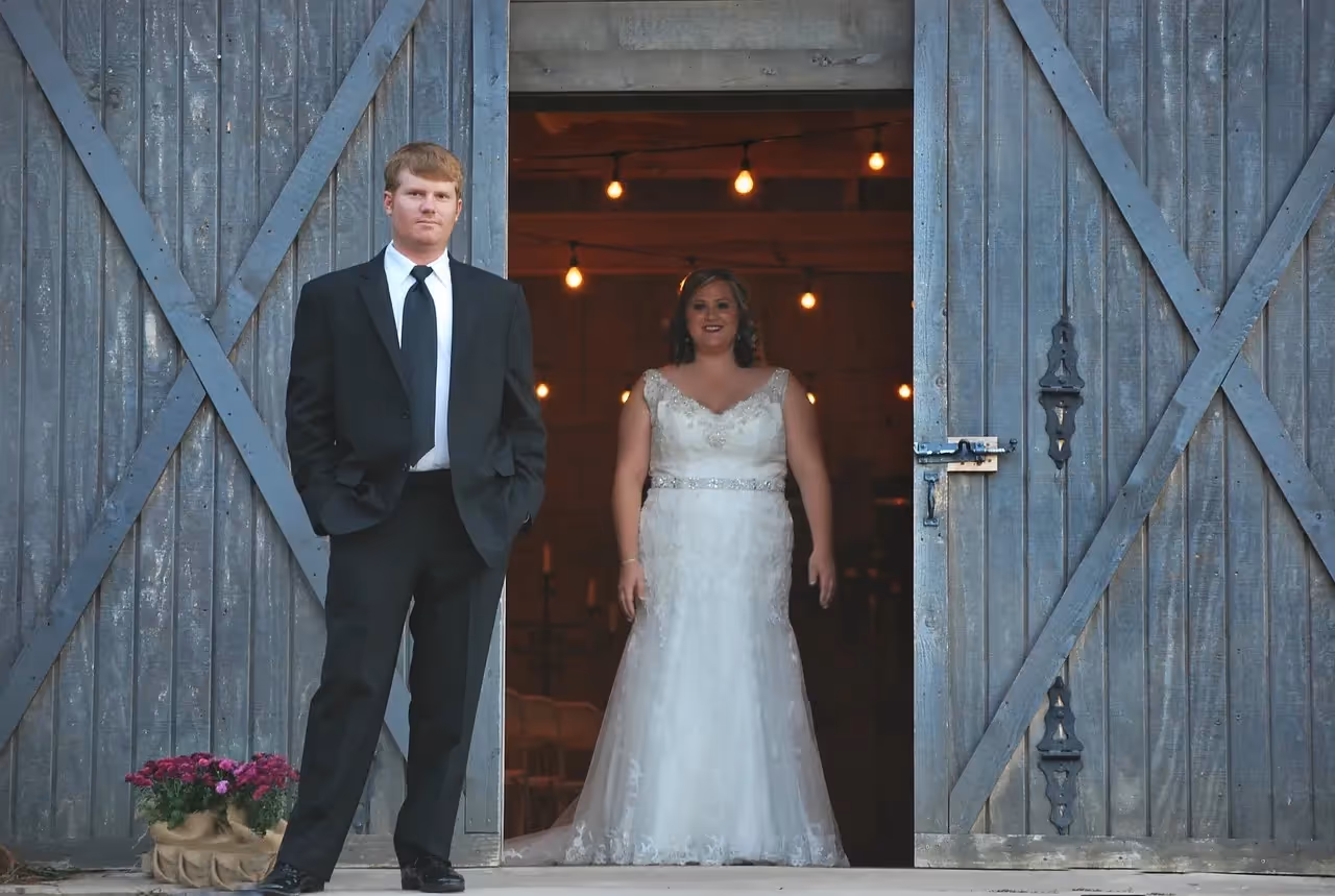 Photo of a groom standing outside barn doors that are half open showing the bride walking up