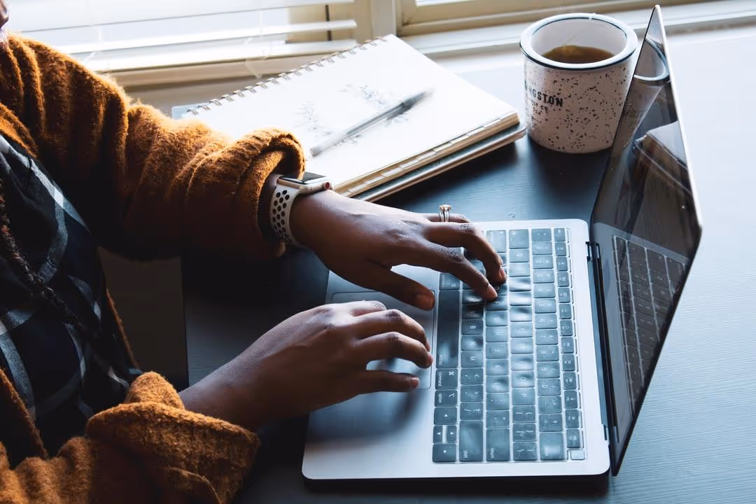 A dark skinned woman sits at a computer with a notebook and a cup of coffee next to her