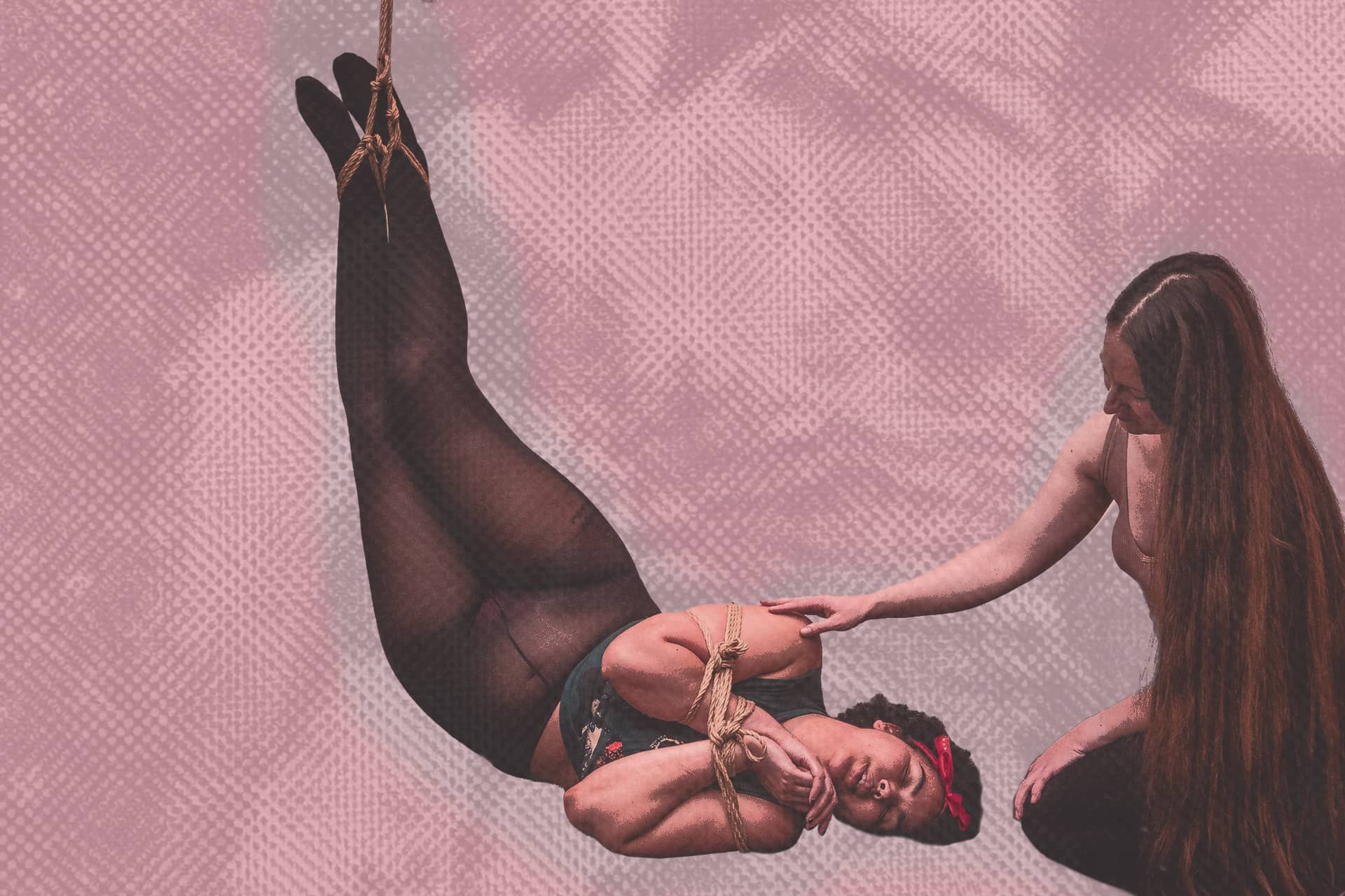 Person suspended in rope harness while another gently supports them against a pink backdrop