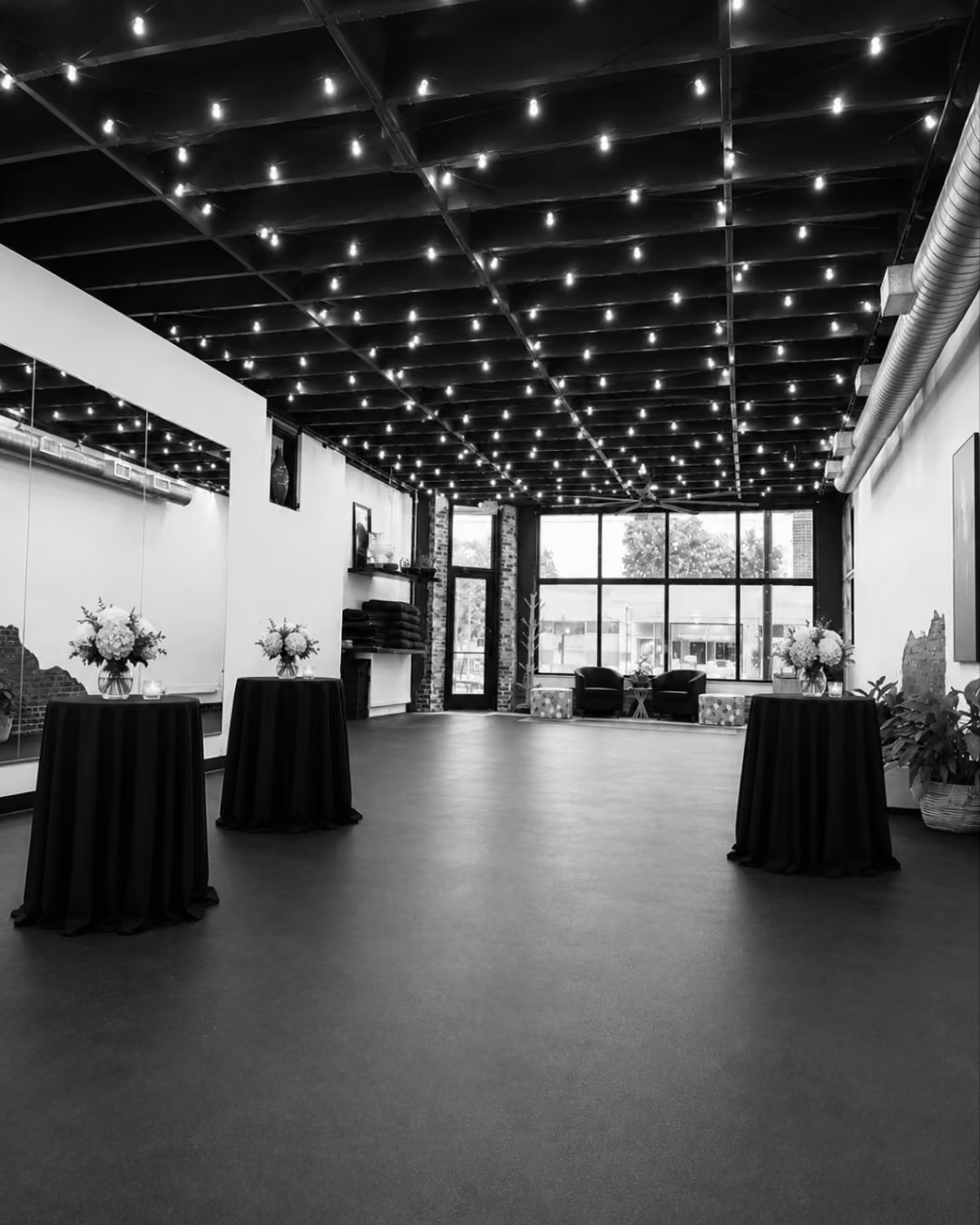 Black and white photo of a spacious indoor venue with string lights on the ceiling, three round cocktail tables covered with dark cloth and floral centerpieces, large windows at the far end, and a mirrored wall with shelves on the left side.