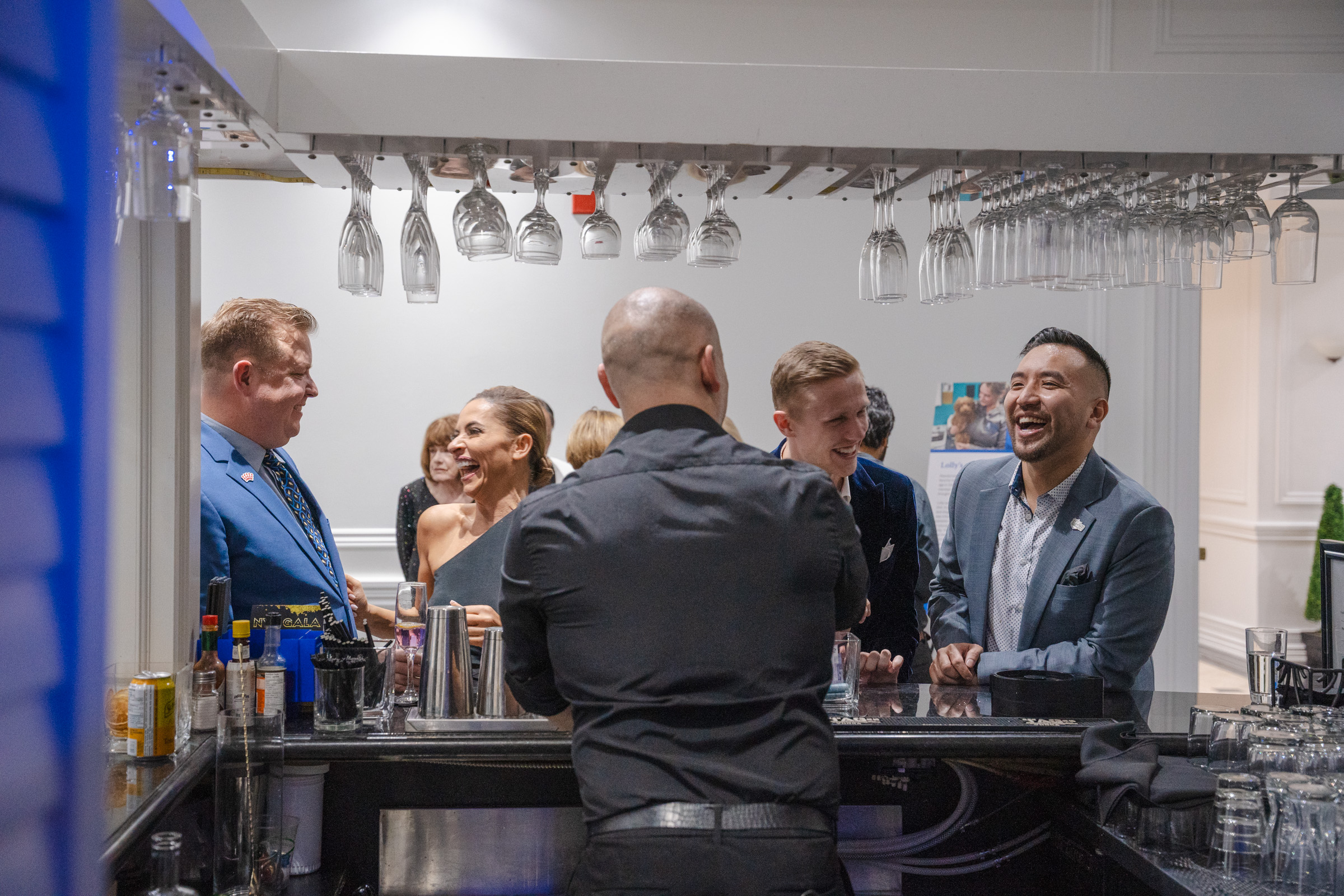 A group of people laugh while standing at a bar while the bartender makes a drink