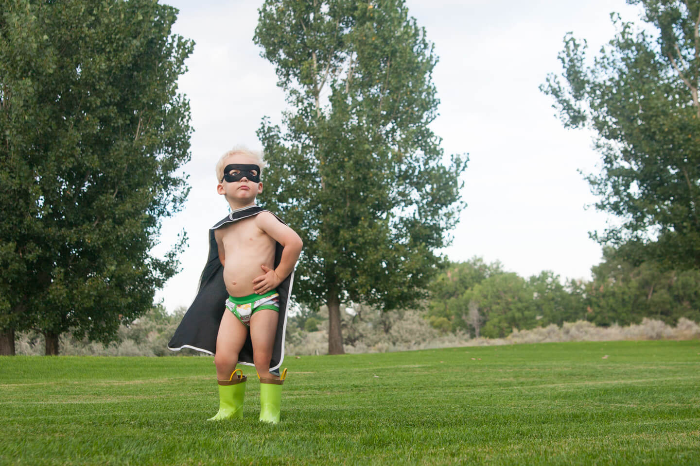 toddler boy stands proudly as a cute little hero in cape and yellow boots outside