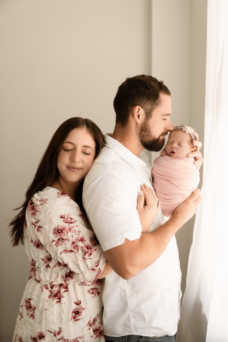 a couple in billings montana kisses their newborn infant daughter. a baby girl sleeps in a studio wicker basket. a cute older sister lovingly snuggles her sleeping newborn brother. best newborn photographer in Billings MT
