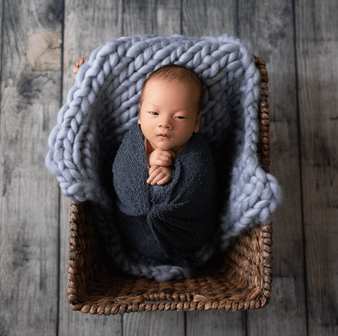 a newborn boy looks straight at the camera in his blue blanket and wicker basket