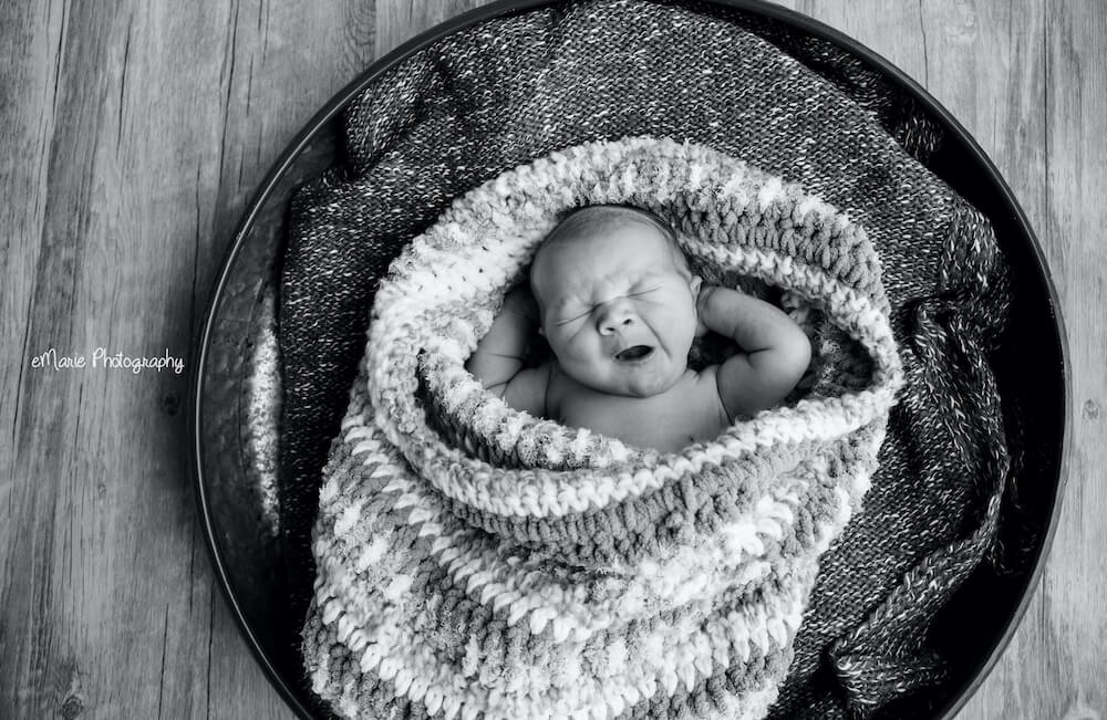 Newborn yawns in a basket while swaddled in a blanket in photography studio