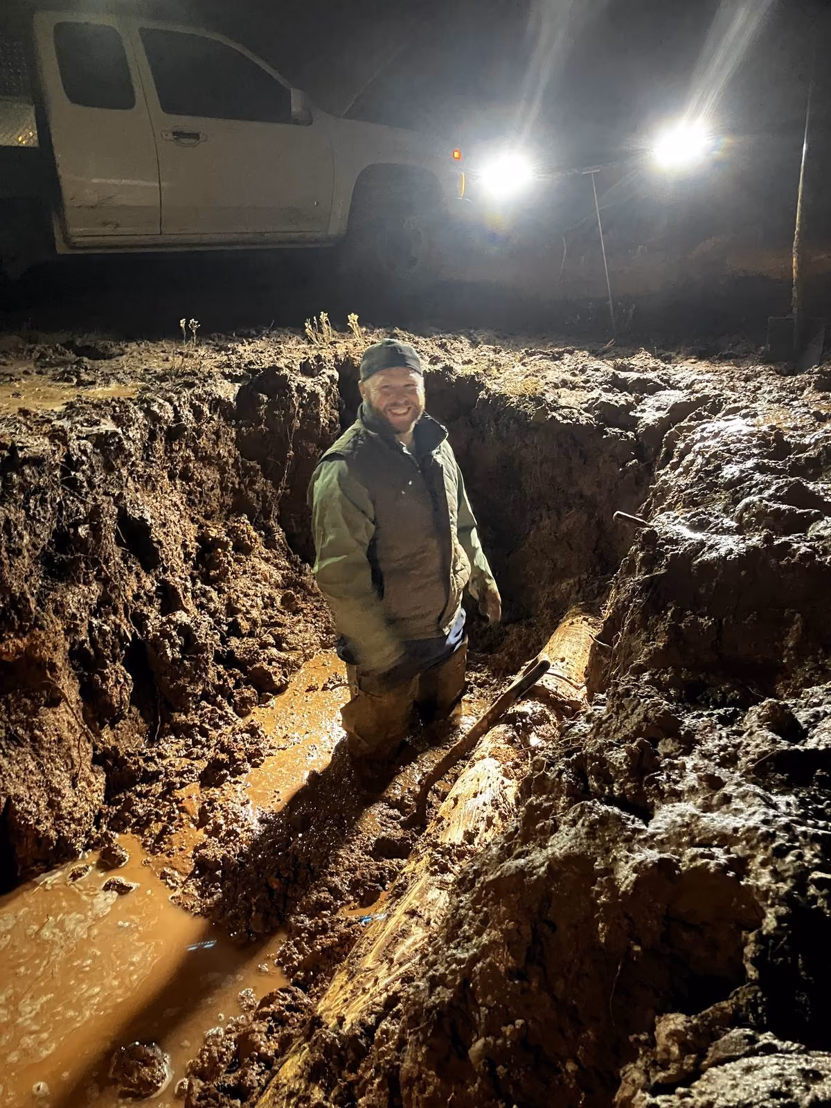 Water utility operator working in muddy excavation trench during nighttime emergency repair