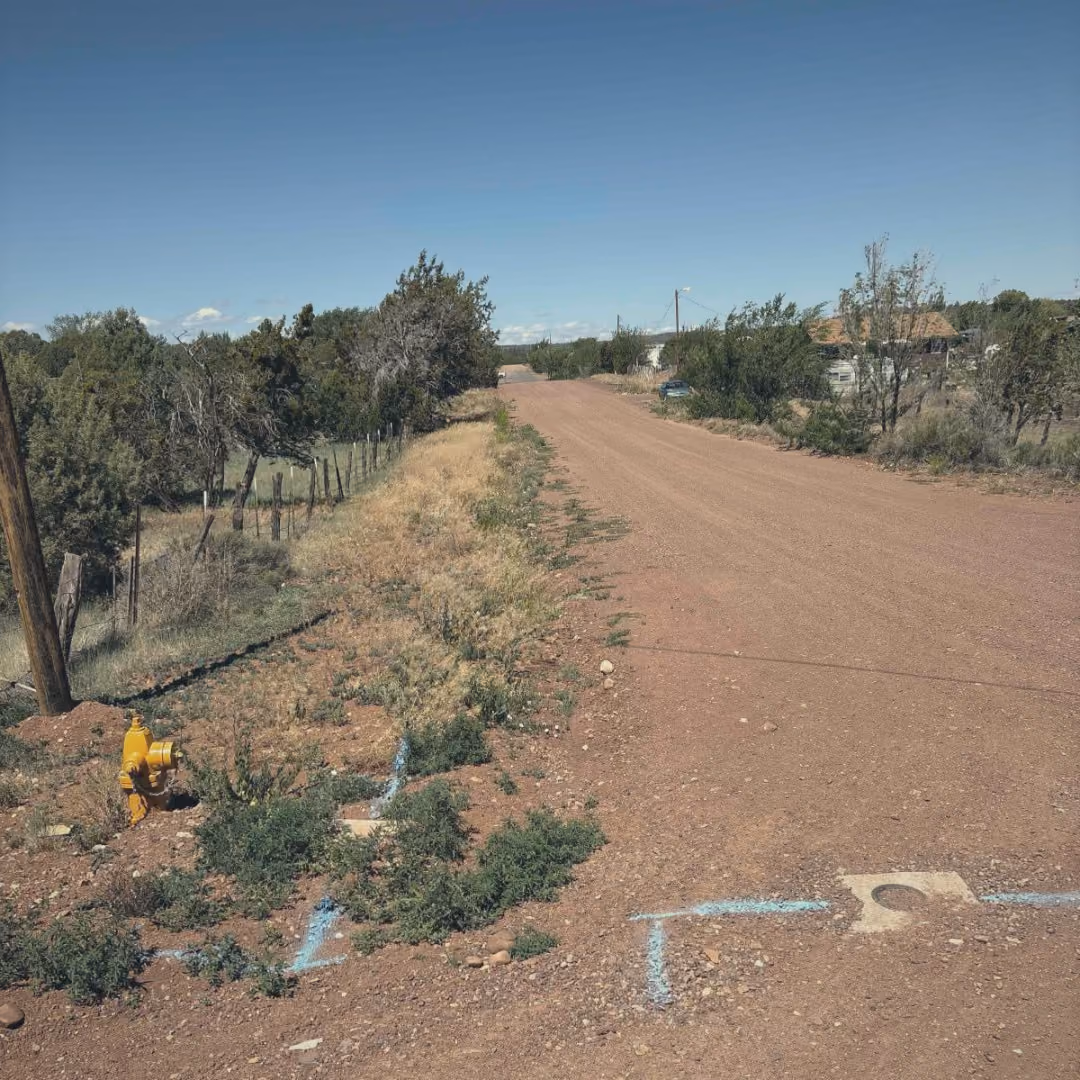 Blue locate paint in a dirt road pointing at a valve box, with a yellow hydrant and fence posts beside it