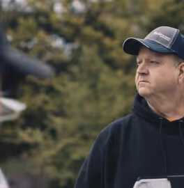 Middle-aged man wearing a black hoodie and blue cap looking to the right with blurred green foliage background.
