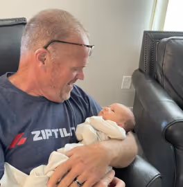 Man wearing glasses and a Ziptilt shirt smiling and holding a newborn baby wrapped in a white blanket while sitting on a black leather chair.
