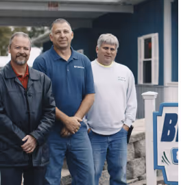 Three middle-aged men standing outdoors beside a building and a sign, casually dressed and smiling.