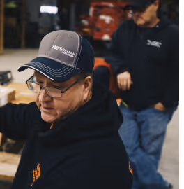 Two men in dark hoodies and caps working in a workshop with wooden materials.