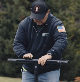 Man wearing a black cap and jacket with an American flag patch working with a tool outdoors.