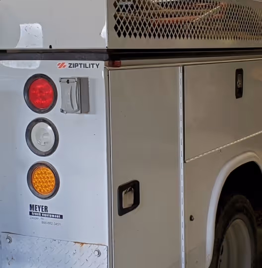 Close-up of the rear side panel of a white utility truck showing brake, reverse, and signal lights, a Ziptility logo, and Meyer truck equipment branding.