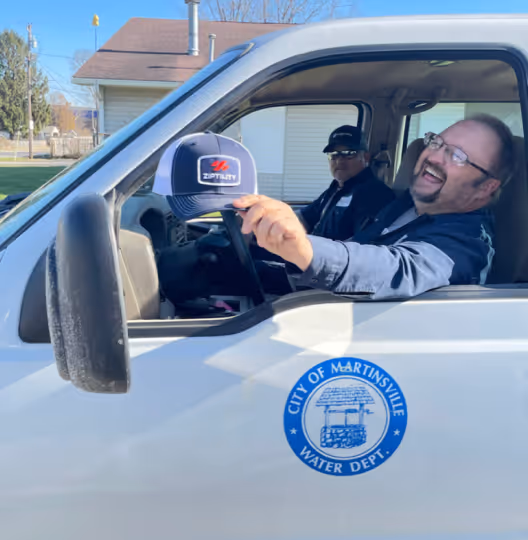 Two men sitting inside a white City of Martinsville Water Department truck, one holding a Ziptility cap out the window smiling.