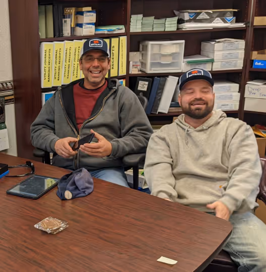 Two men wearing caps and casual hoodies sit at a wooden table in an office filled with shelves and files, smiling.