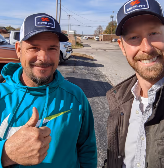 Two men outdoors on a sunny day, both wearing navy blue Ziptivity caps; one man in a teal hoodie giving a thumbs-up, the other in a brown jacket smiling.