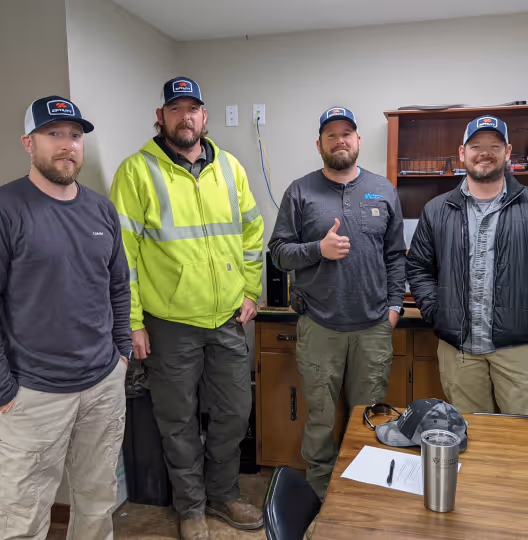 Four men in casual and work clothing wearing matching hats standing indoors near a wooden table with office items.