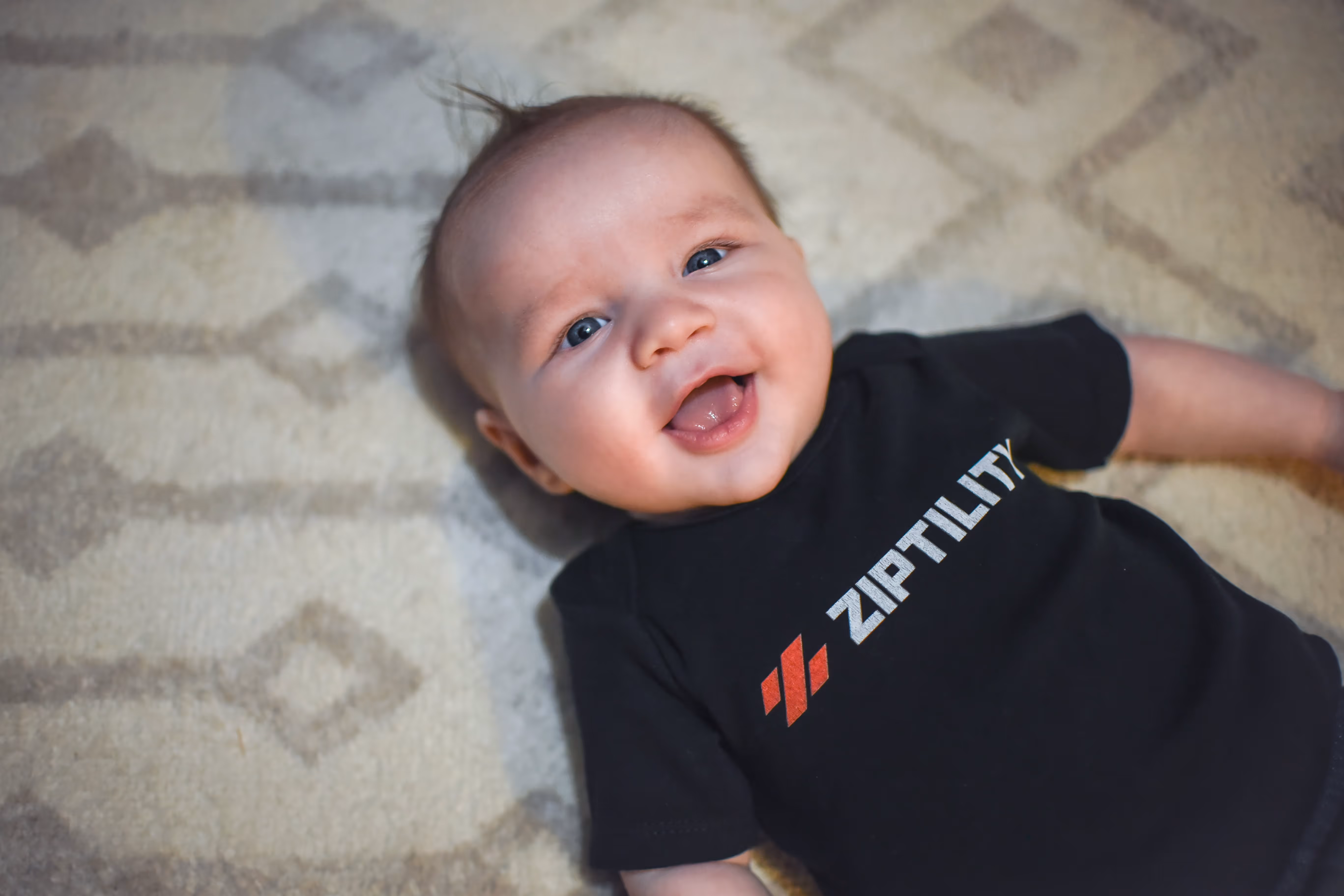 Smiling baby lying on patterned carpet wearing a black Ziptility t-shirt.