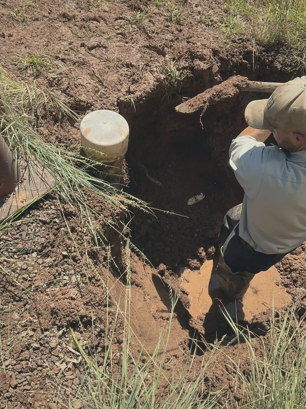 Operator standing in a muddy hand-dug excavation beside a capped riser, getting eyes on what's in the ground.