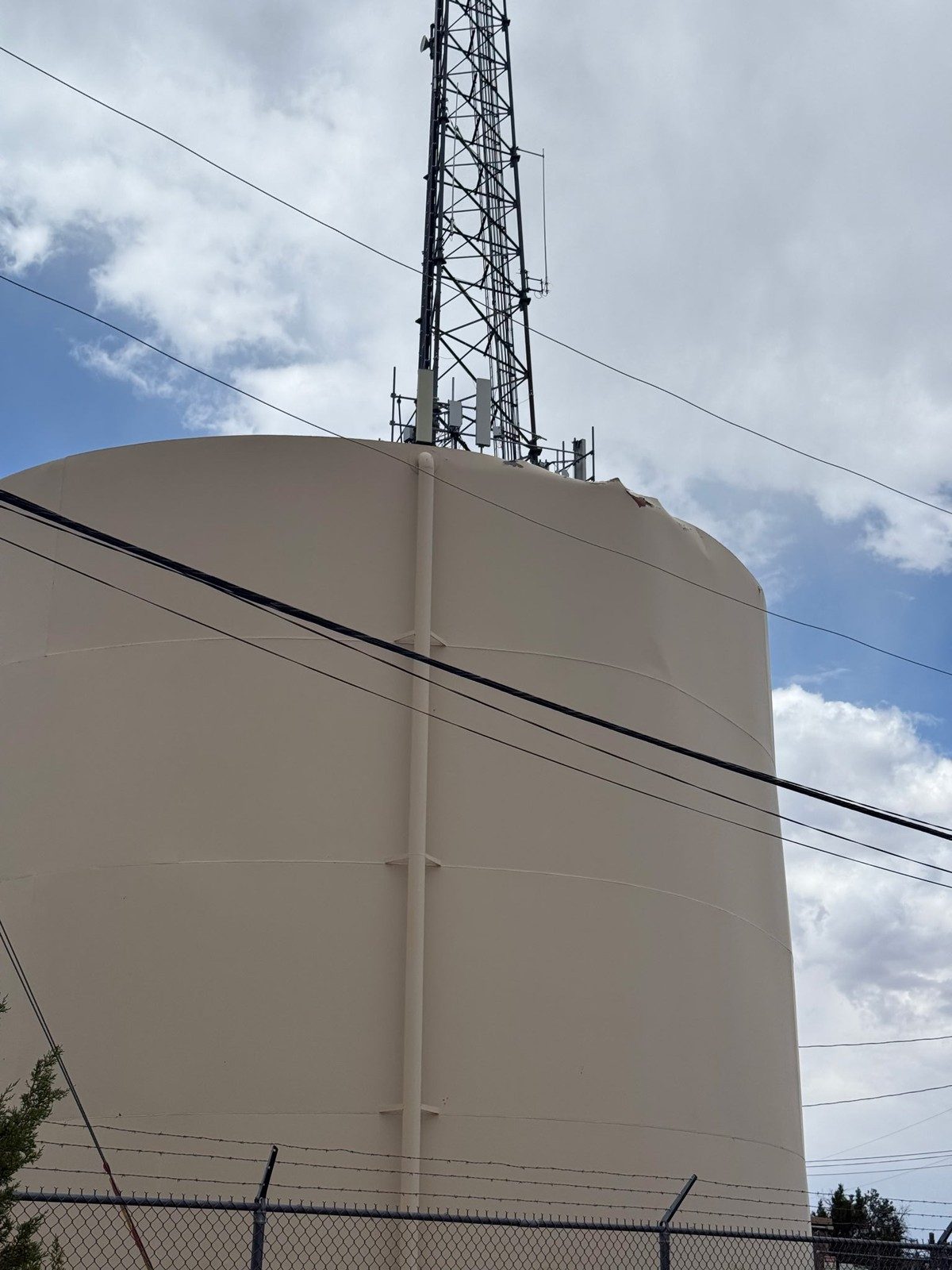 The Heber DWID storage tank seen from outside its fence after the failure: a tall tan welded steel tank with a cell tower on the roof, the roof edge torn at the upper right.