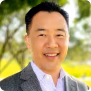 Smiling man in a gray blazer and white shirt with green trees in the background.