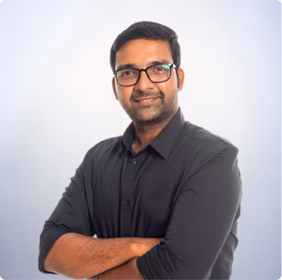Smiling man with glasses and black shirt stands with arms crossed against light background.