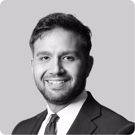 Smiling man with short hair and beard wearing a dark suit and tie on a gray background.