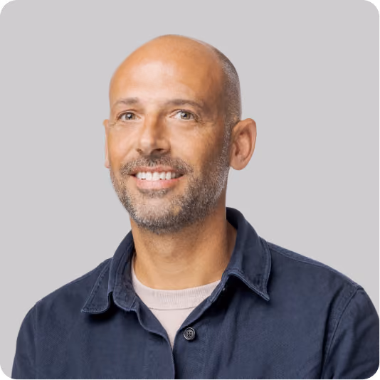 Smiling bald man with short beard wearing a dark jacket over a light shirt against a gray background.