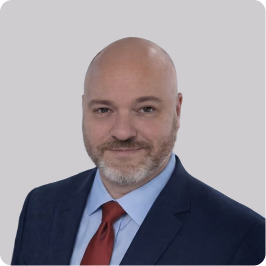 Smiling bald man with gray beard wearing a navy suit, blue shirt, and red tie against a gray background.
