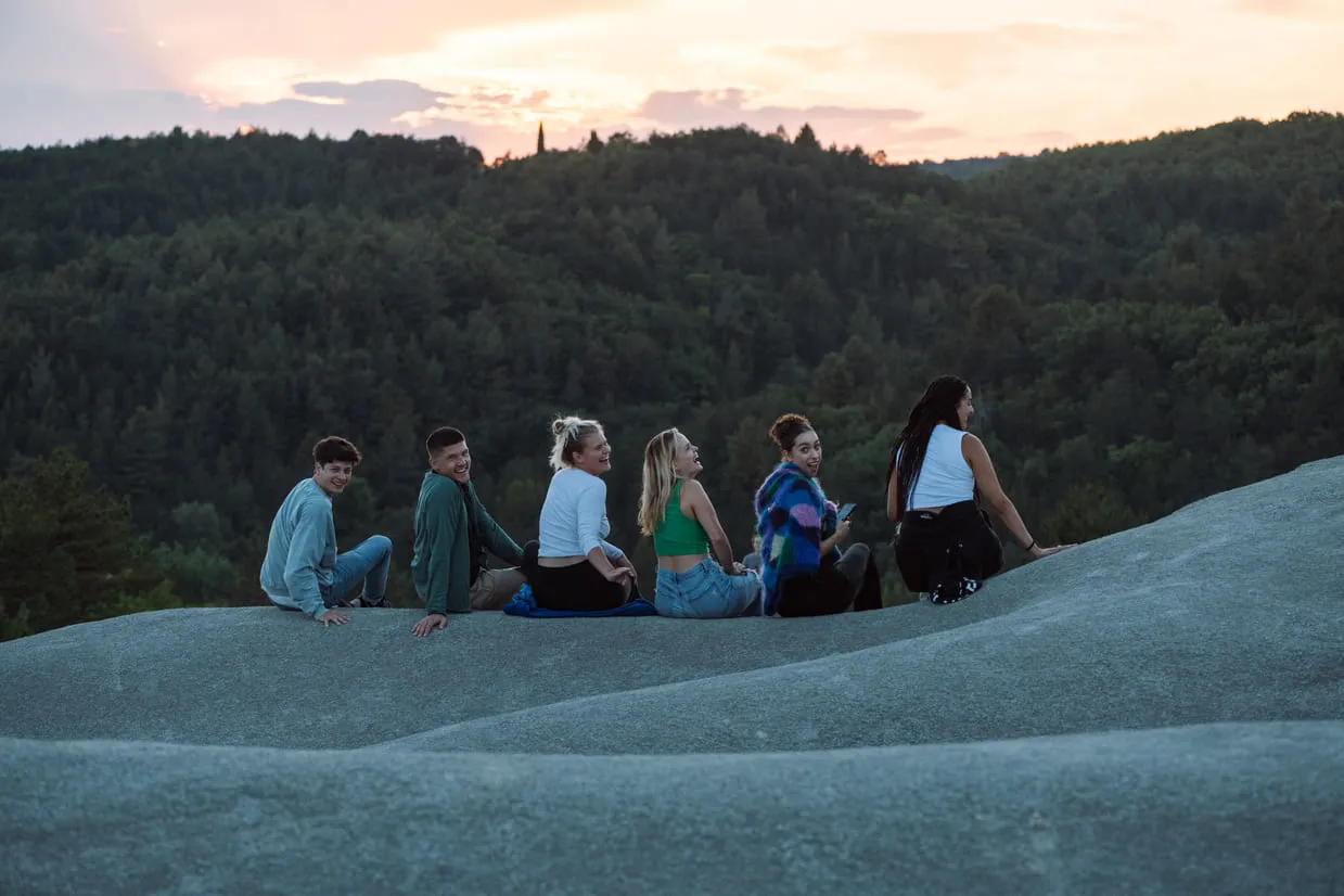 Sechs junge Menschen sitzen nebeneinander auf einer Anhöhe, einige schauen zur Kamera, dahinter Wald und Abendhimmel.