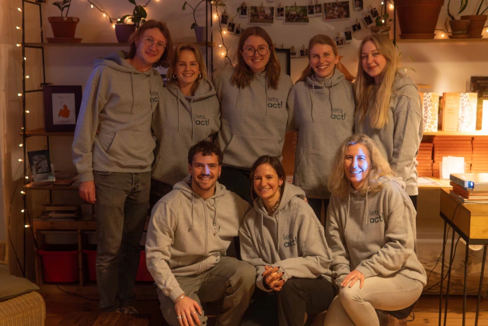 Acht lächelnde Erwachsene in grauen Kapuzenpullovern mit 'lets act!' sitzen und stehen in einem gemütlich beleuchteten Raum mit Pflanzen und Fotos an der Wand.