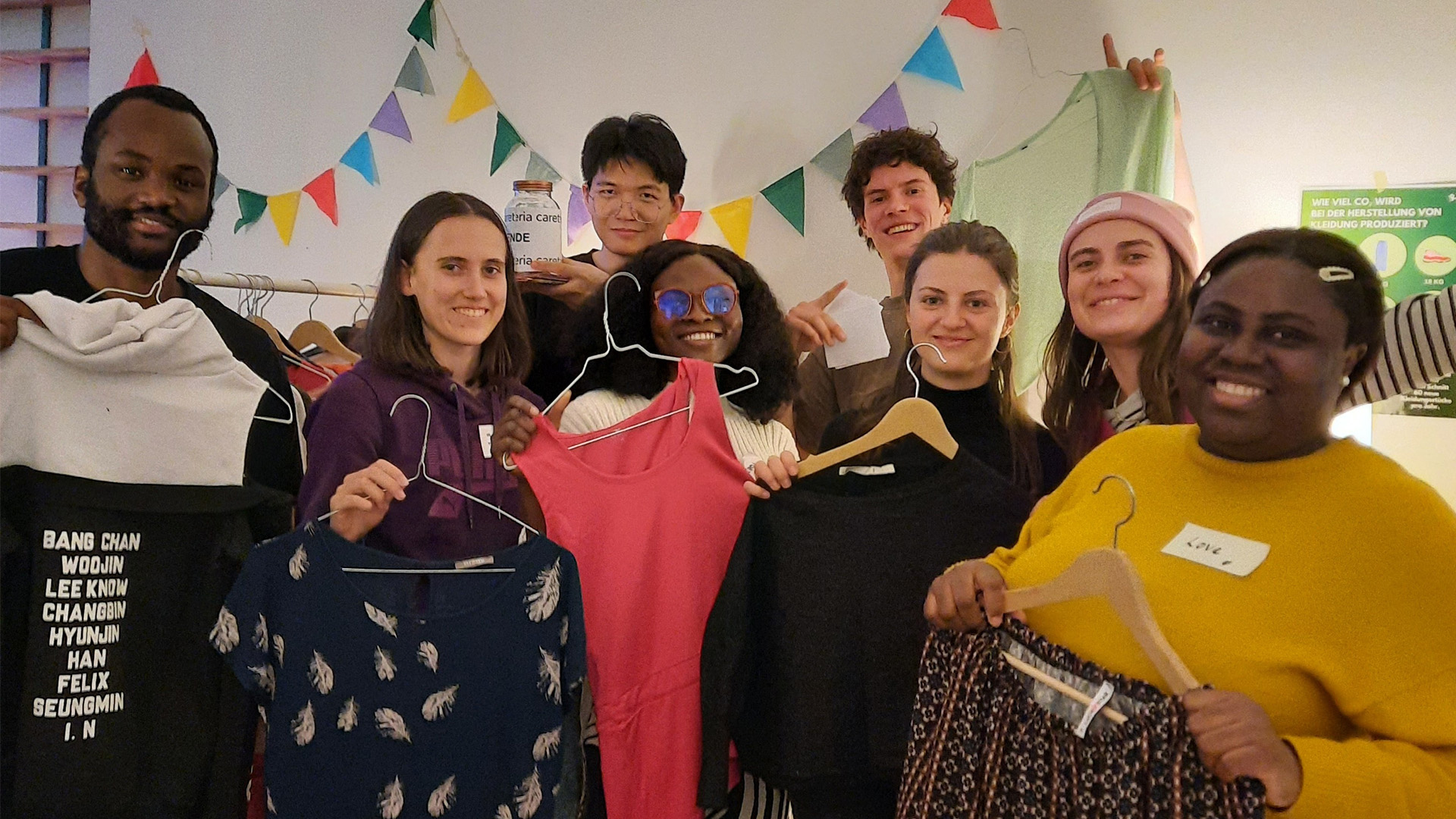 Acht Personen stehen lachend vor einer weißen Wand mit bunten Wimpeln. Sie halten bunte Kleidungsstücke auf Bügeln in die Kamera: eine dunkelblaue Bluse mit Feder-Muster, ein korallenrotes Top und ein gemustertes dunkelbraunes Kleidungsstück. Ein Mann trägt ein K-Pop-Band-T-Shirt (mit Namen wie „Bang Chan" sichtbar), eine Person trägt eine rosarot Wollmütze und blaue Sonnenbrille. Die Gruppe wirkt fröhlich und verbunden, zusammen bei einer Kleidertauschparty.