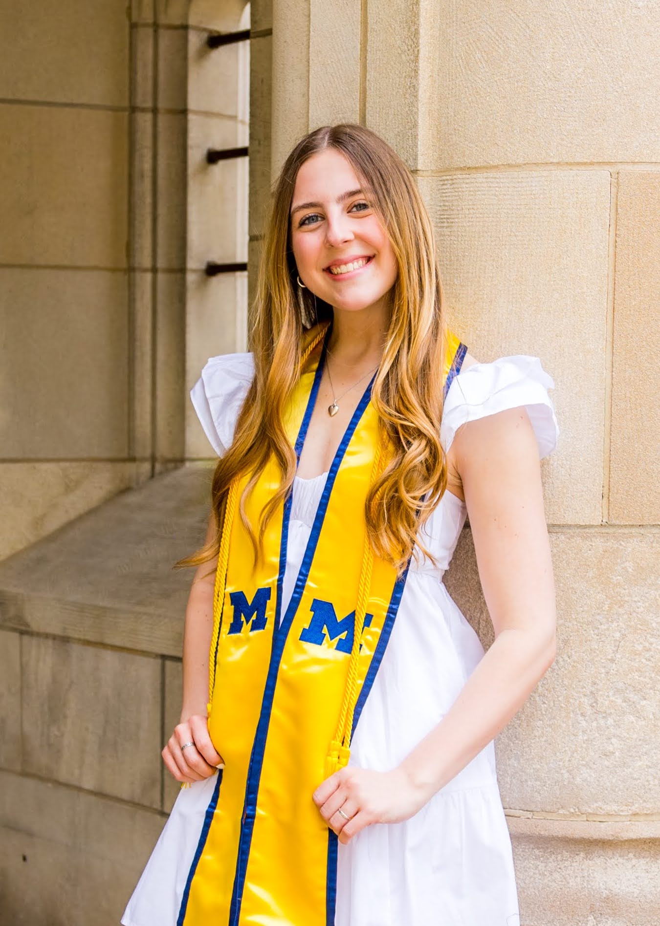 Brunette girl in white dress and yellow University of Michigan sash leaning against a building column
